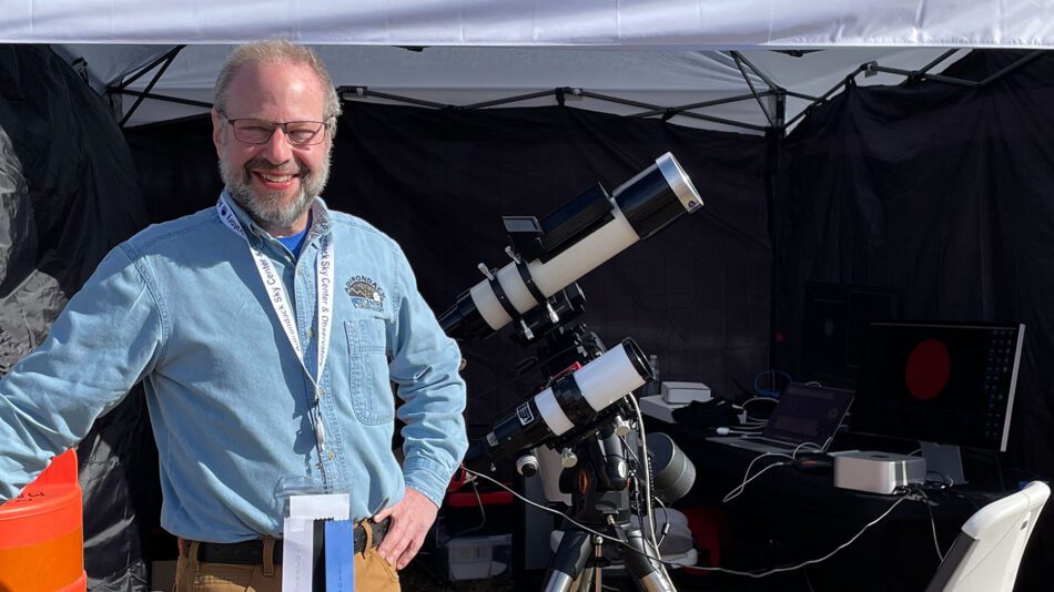 smiling man with telescope and NASA live feed tent behind him