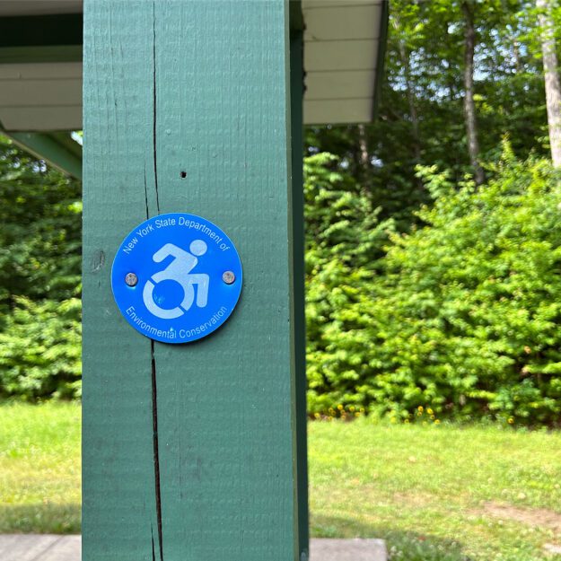 Green wooden pillar of a campsite bathroom with a wheelchair-accessible medallion on it.