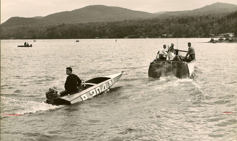 Black and white photo of a race boat being towed by another boat on a lake.