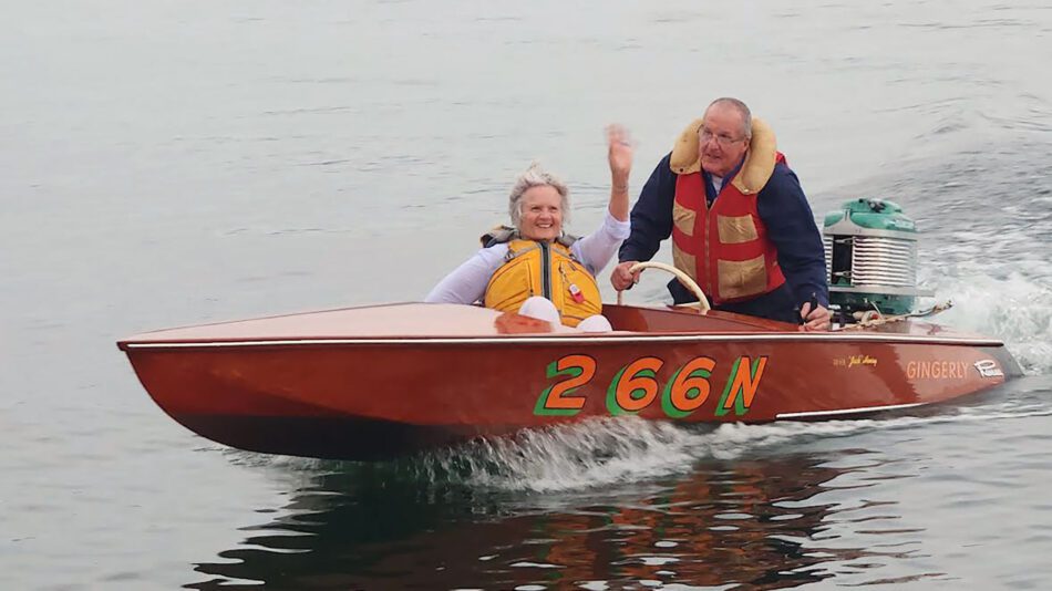 Woman waves from the front of a small wooden boat on the water. She is wearing an orange life jacket. A man in a red life jacket sits behind her and drives the boat.