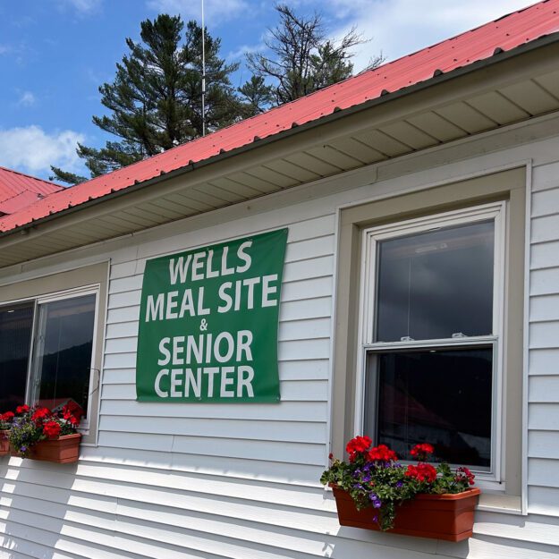 Outside of a white building with flower boxes and a sign that says "Wells Meal Site and Senior Center".