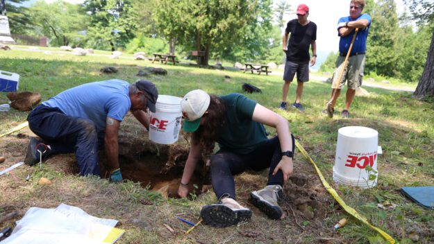 People dig in a test pit at the Lake George Battlefield while two bystanders watch