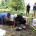 People dig in a test pit at the Lake George Battlefield while two bystanders watch