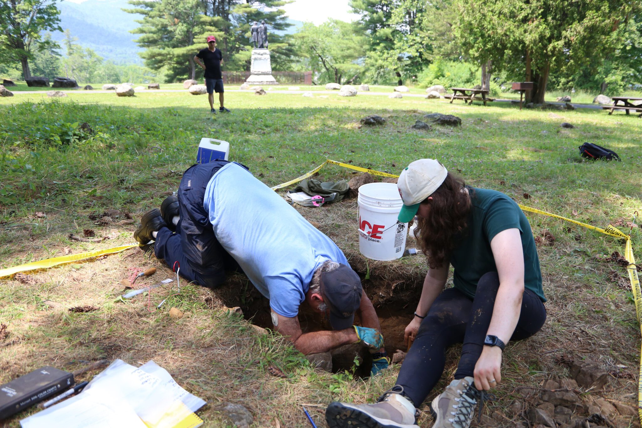 A bystander watches two people digging at the Lake George Battlefield