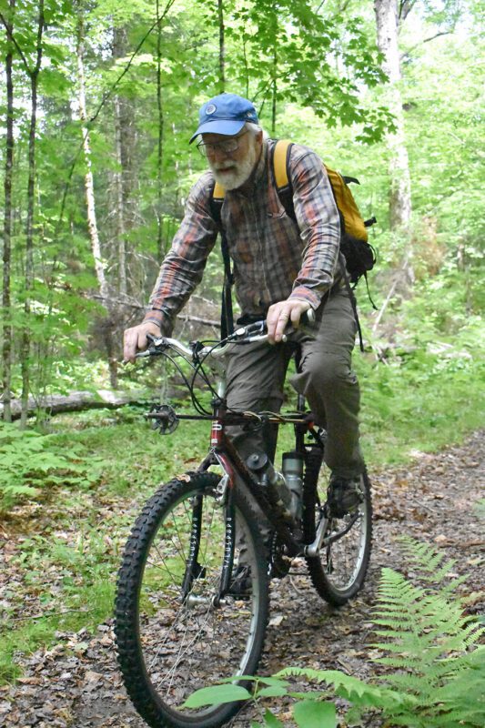 A man in a plaid shirt with a blue hat rides a bike in the woods.