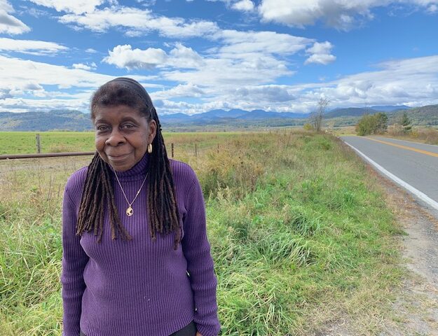 Author Alice Green, wearing purple and standing near green plants at a roadside