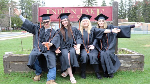 Four students in black caps and gowns sit together in front of their high school's sign.