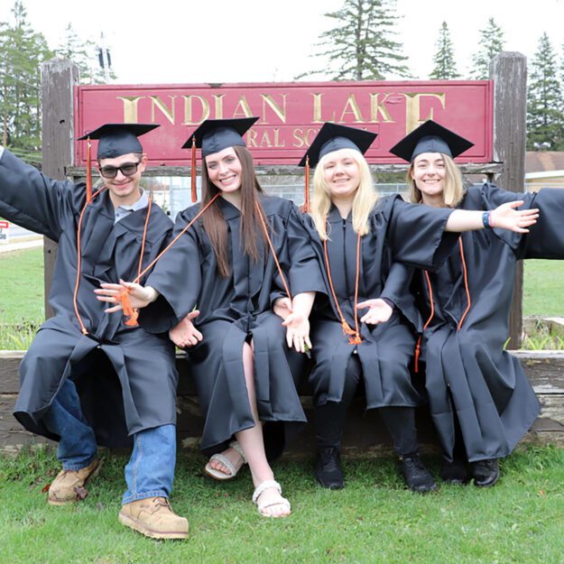 Four students in black caps and gowns sit together in front of their high school's sign.