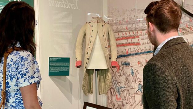 Two museum visitors look at a faded white and pink vintage army uniform.