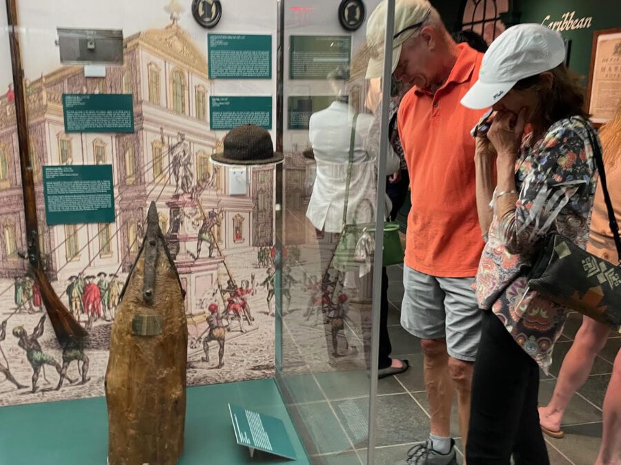 Two museum visitors look at war artifacts, including a gun and brown hat, through a glass box.