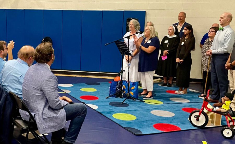 woman standing at a microphone along with other people standing on a multi-colored rug addressing a seated audience